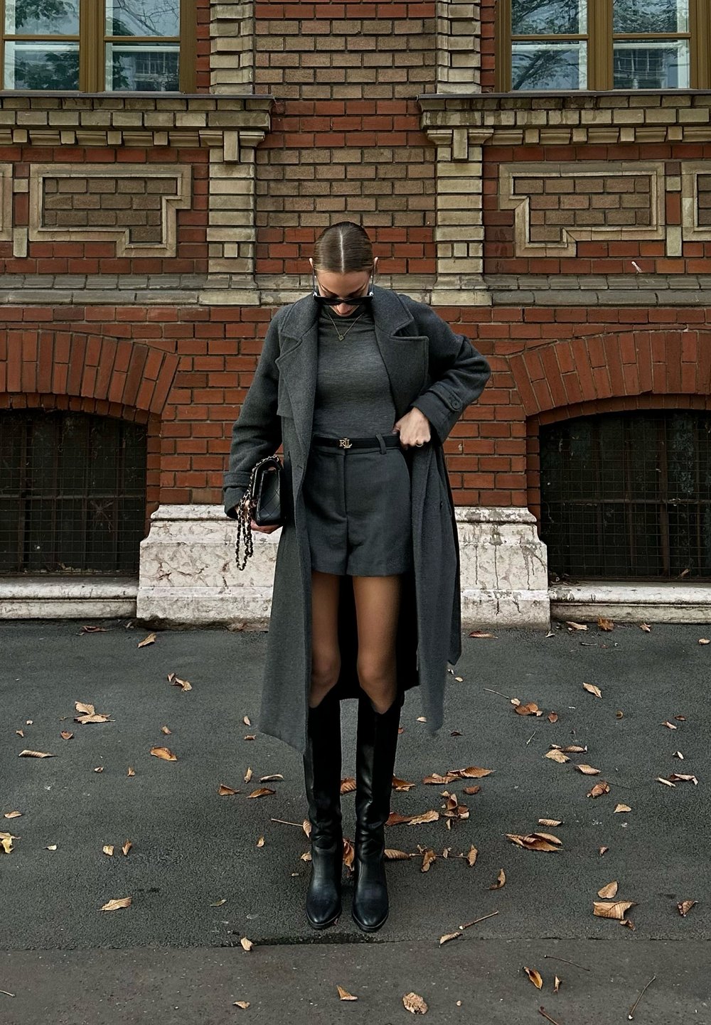 Un manteau en laine gris porté par-dessus un pull gris ajusté et un short taillé. Des bottes noires jusqu'aux genoux et un petit sac à main noir avec une chaîne. Fond en briques brunes.