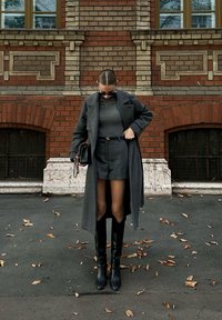 Un manteau en laine gris porté par-dessus un pull gris ajusté et un short taillé. Des bottes noires jusqu'aux genoux et un petit sac à main noir avec une chaîne. Fond en briques brunes.