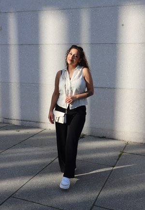 Blusa sin mangas de color gris claro, pantalones de pierna ancha negros, zapatillas blancas y un bolso cruzado crema con un cierre dorado contra una pared texturizada.