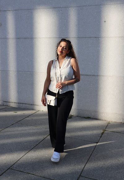 Blusa sin mangas de color gris claro, pantalones de pierna ancha negros, zapatillas blancas y un bolso cruzado crema con un cierre dorado contra una pared texturizada.