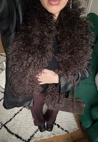 Personne portant une veste en cuir noire avec un grand col en fourrure bouclée, un pantalon bordeaux, des chaussures noires, et tenant un sac à main en daim marron.