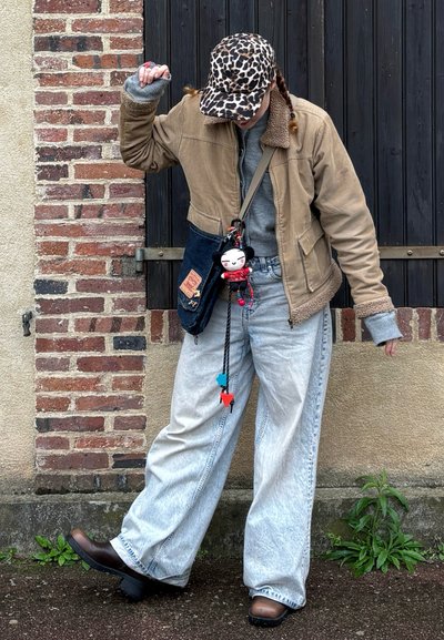 Chaqueta de pana marrón con un cuello de pelaje, sudadera gris, jeans holgados de un azul claro, botas de tacón marrones. El accesorio incluye un bolso con un charm de personaje.