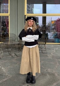 Svart puffermet topp over en hvit skjorte med krage, beige knelangt skjørt og svarte kraftige ankelstøvler. Svart beret med logodetalj.