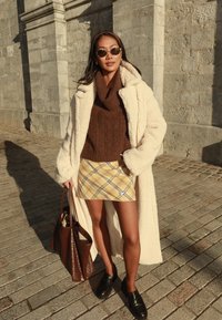 Kreemjas fliisist pikk mantel; pruun kootud kampsun; kollane ruuduline miniseelik; mustad loafers-tüüpi kingad; pruun käekott; ümmargused päikeseprillid. Kivist taust.