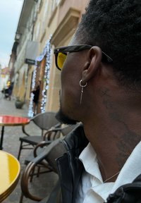 Schwarze Sonnenbrille mit gelben Gläsern, silberner Ringohrring mit einem hängenden Akzent und ein sichtbares Tattoo am Hals. Außenbereich eines Cafés.