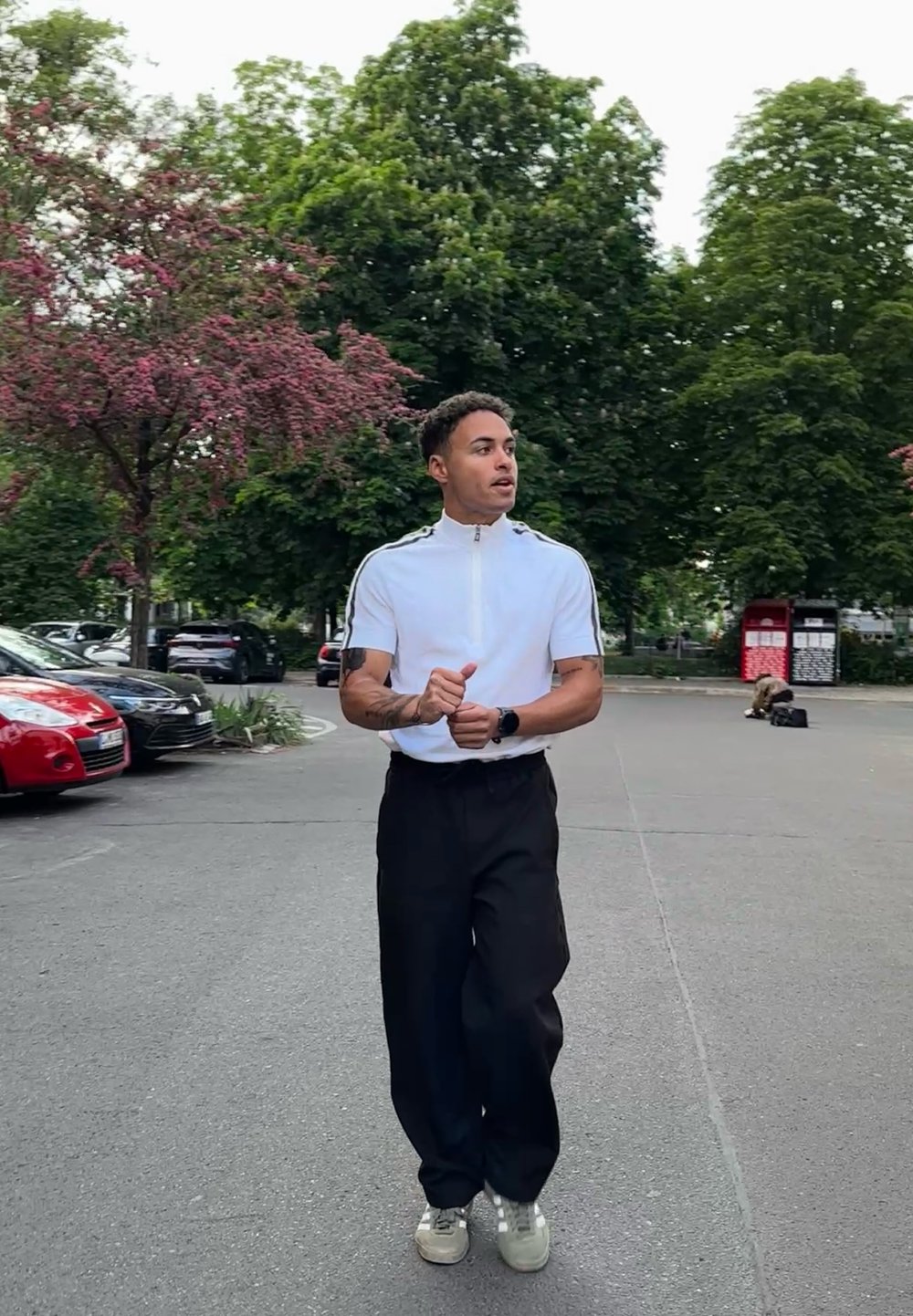 Weißes Reißverschluss-Polohemd mit schwarzen seitlichen Streifen, schwarze weite Hosen und hellgraue Sneakers; im Hintergrund sind grüne Bäume und geparkte Autos zu sehen.