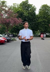 Weißes Reißverschluss-Polohemd mit schwarzen seitlichen Streifen, schwarze weite Hosen und hellgraue Sneakers; im Hintergrund sind grüne Bäume und geparkte Autos zu sehen.