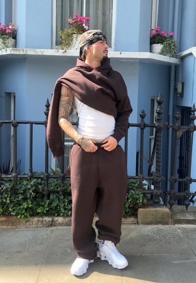 Sudadera marrón colocada sobre una camiseta sin mangas blanca, combinada con pantalones de chándal marrones a juego y zapatillas blancas. Tatuajes notables en el brazo.