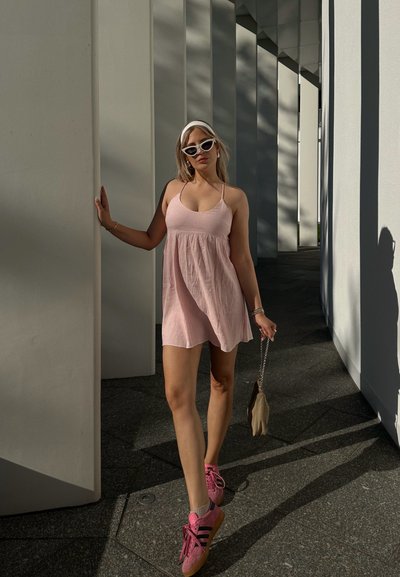 Mujer con un vestido rosa pálido y zapatillas rosas se apoya en un pilar, usando gafas de sol blancas con forma de ojo de gato y sosteniendo un pequeño bolso beige.