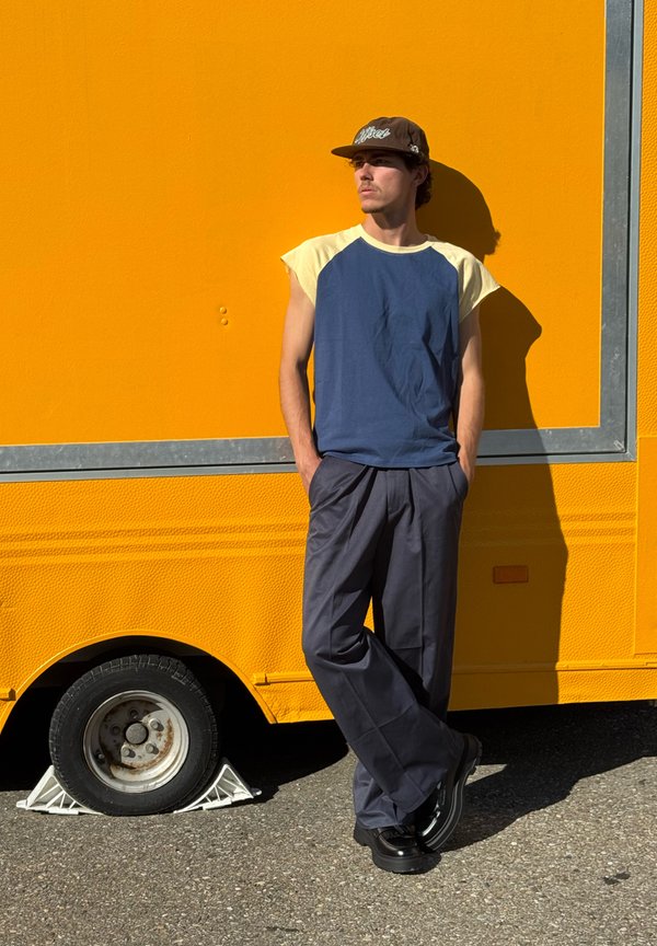 Young man wearing a cap and casual clothes leans against a bright yellow vehicle with hands in pockets, standing on asphalt under sunlight.