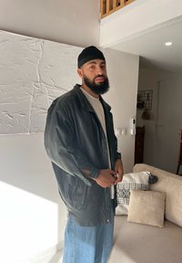 Μαύρο δερμάτινο oversized μπουφάν με κλείσιμο με κουμπί, φορεμένο πάνω από άσπρο πουκάμισο. Συνδυάζεται με μπλε παντελόνι jeans σε φαρδιά γραμμή. Ελαφρώς υφασμένη φόντο.