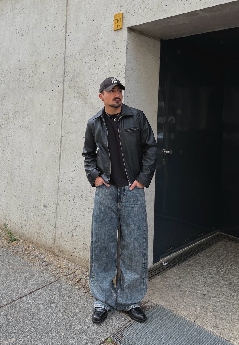Homme portant une veste en cuir noire, un jean bleu ample, des chaussures noires et une casquette noire, debout contre un mur en béton près d'une porte sombre.