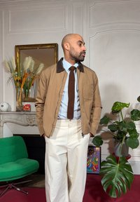 Veste beige avec col marron foncé, chemise bleu clair et cravate marron texturée. Pantalon blanc avec une poche zippée. Fauteuil vert en arrière-plan.