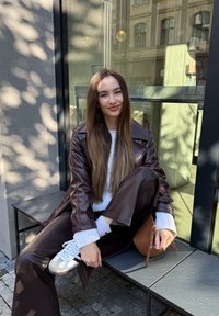 Mlada ženska v rjavi usnjeni obleki in belem puloverju sedi na klopi pred stavbo ter drži majhno rjavo torbico.