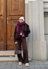 Femme portant un bonnet en tricot blanc, une écharpe bordeaux, une veste en fourrure foncée, un pantalon marron, tenant un petit sac à main sombre, debout près d'une porte en bois et d'un mur en pierre.