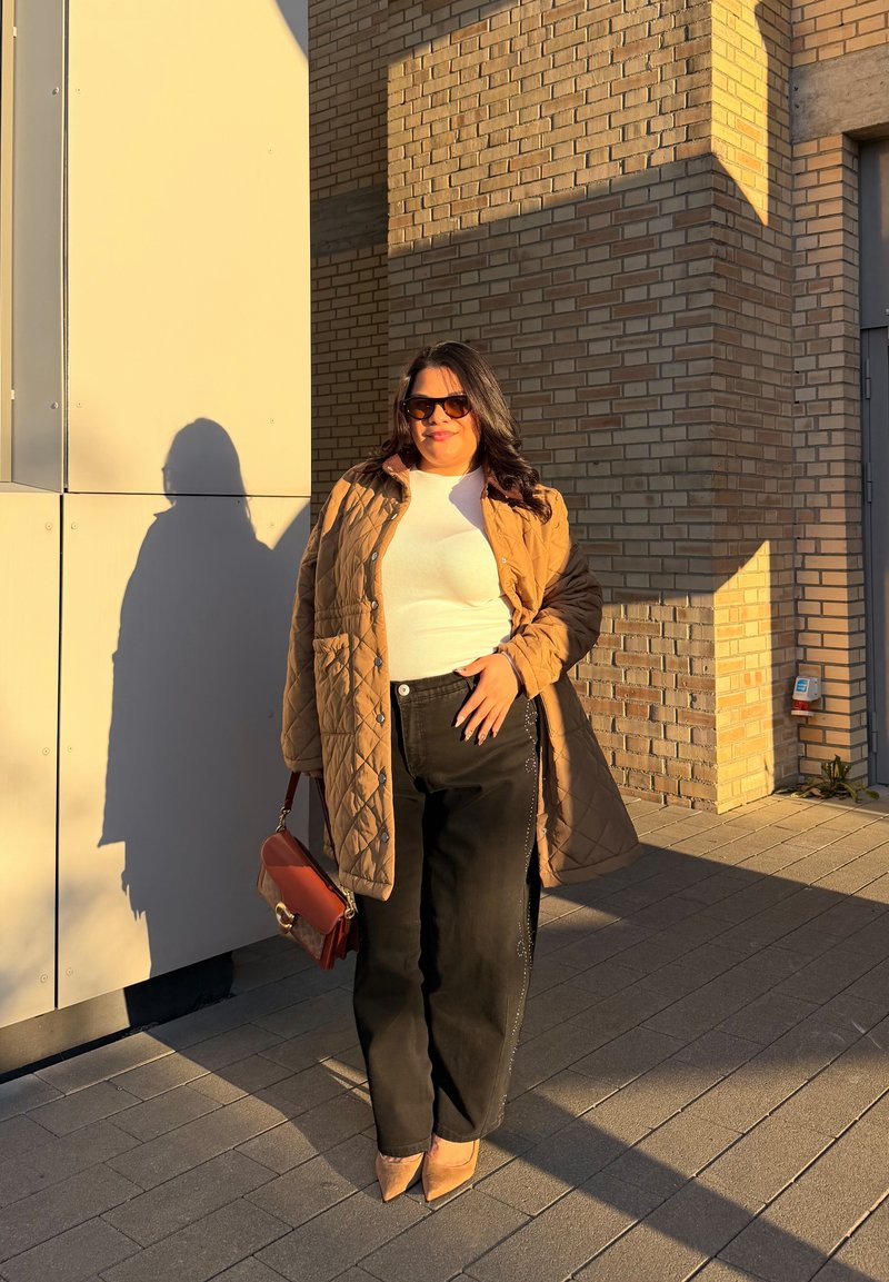 Femme portant des lunettes de soleil, manteau matelassé beige, haut blanc, pantalon noir, talons beiges, tenant un sac à main marron, debout près d'un mur en briques au soleil.