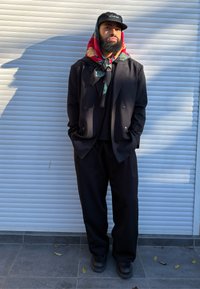 Giacca doppiopetto nera, pantaloni neri larghi, scarpe nere, sciarpa multicolore con motivi floreali e paisley, berretto da baseball.