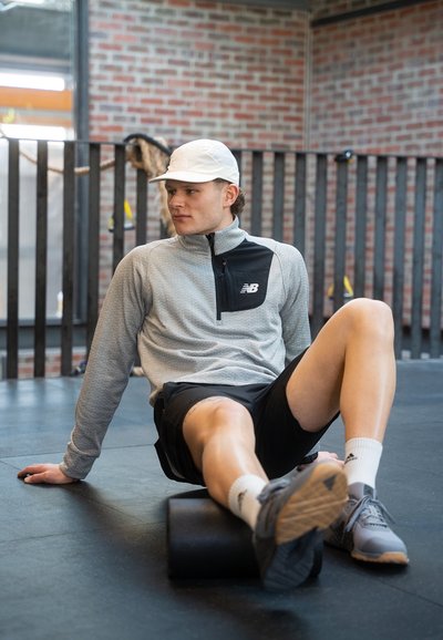 Chaqueta gris con cremallera y panel frontal negro, rodillo de espuma negro, pantalones cortos deportivos, zapatillas gris con suelas texturizadas y una gorra blanca.