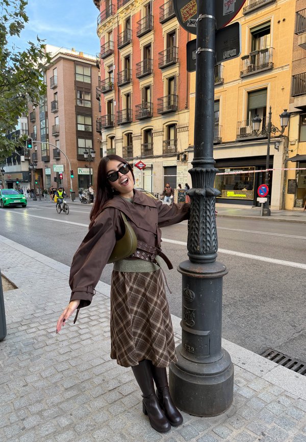 Une femme portant des lunettes de soleil pose à côté d’un réverbère orné sur un trottoir en ville. À l’arrière-plan, on aperçoit des bâtiments historiques, des piétons, un cycliste et la circulation.