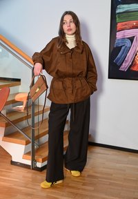 Brun, oversizejacka med dragsko, kombinerad med svarta byxor med vida ben och gula skor. Modellen håller en brun handväska med gulddetalj.