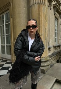 Musta toppatakki hupulla, päällä valkoinen t-paita. Yhdistetty maastokuvioisiin shortseihin ja pörröiseen mustaan laukkuun. Tyylikkäät aurinkolasit viimeistelevät asun.