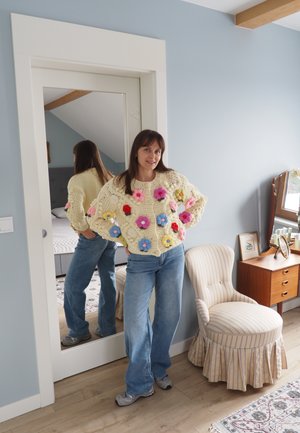 Mujer de pie con las manos en las caderas, vistiendo un suéter crema con flores de ganchillo coloridas, jeans azules y zapatillas grises junto a un espejo.