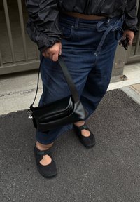 Borsa a mano in pelle nera con forma curva e una sola tracolla, tenuta in una mano. Abbinata a pantaloni in denim blu scuro larghi e calzature nere.