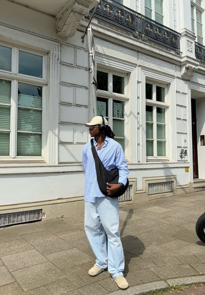 Camisa de botones a rayas azul claro, jeans de mezclilla livianos y holgados, sombrero beige, bolso bandolera negro, auriculares negros, de pie cerca de un edificio blanco.