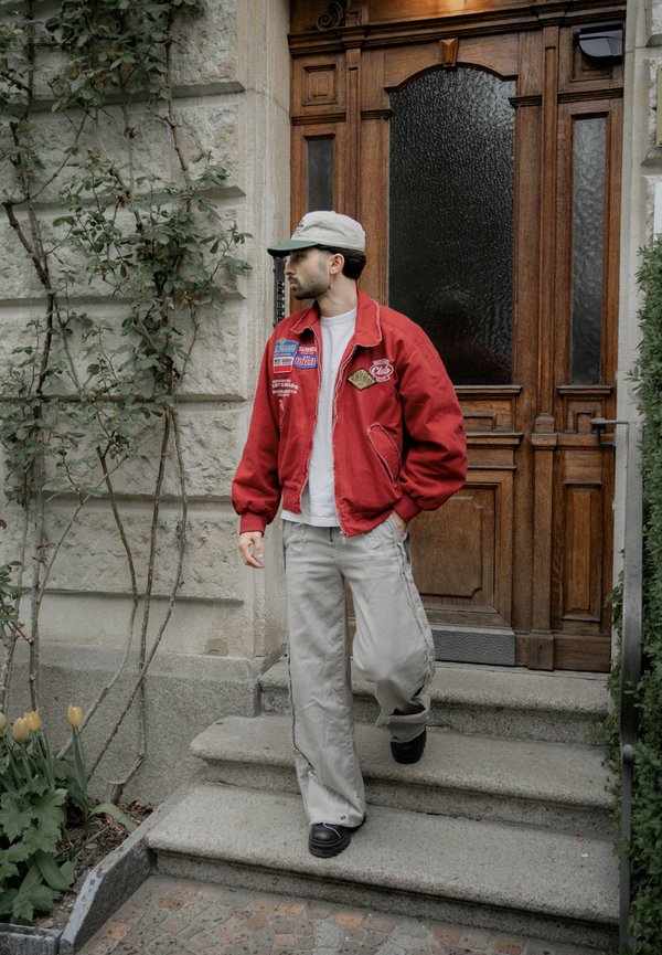 Mladý muž v červenej bunde a šedej čiapke schádza po kamenných schodoch pred drevenými dverami s rastlinami v blízkosti.