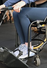 Person iført blå leggings og joggesko bruker en romaskin mens de sitter i en rullestol på et treningsstudio.