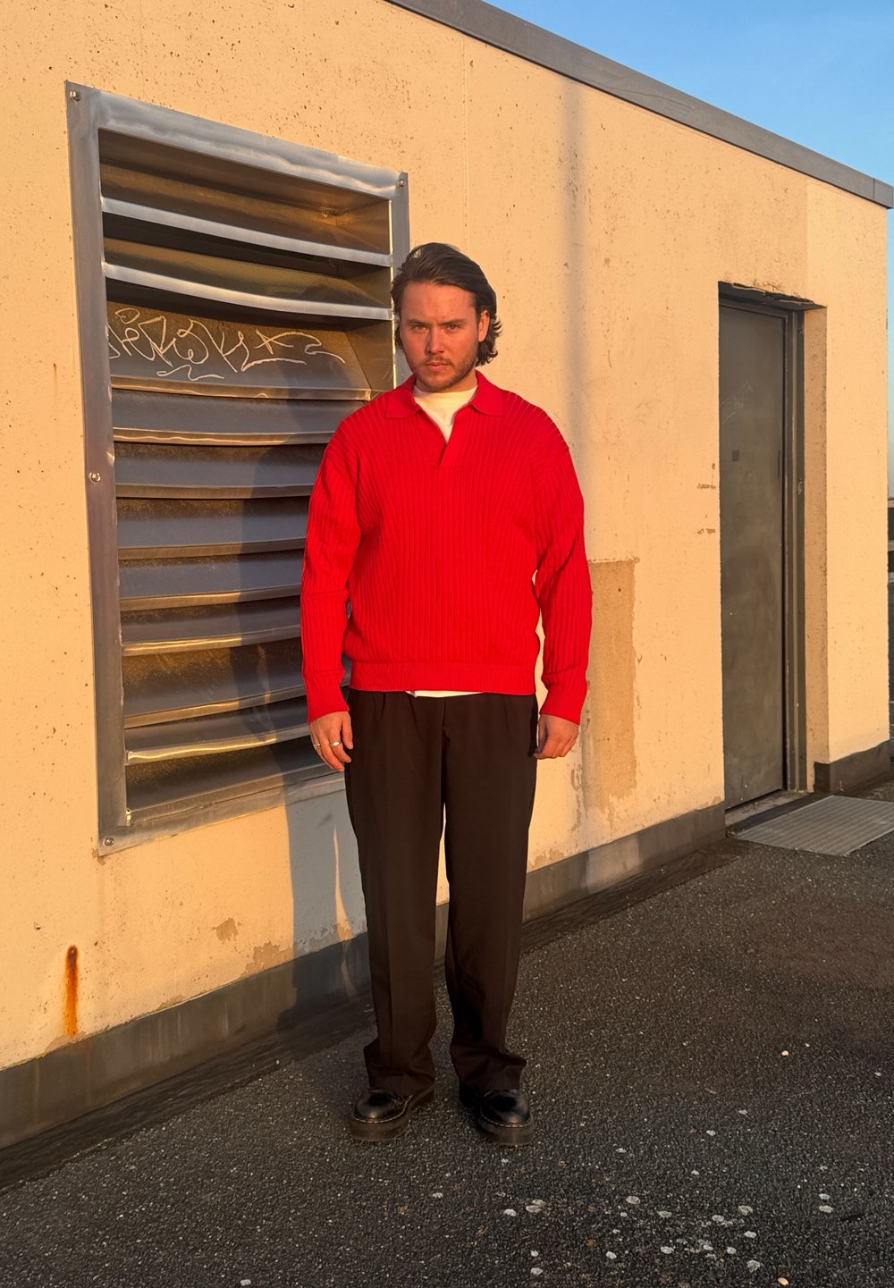 Κόκκινο ριπμένο πλεκτό πουλόβερ με γιακά, φορεμένο πάνω από λευκό πουκάμισο; μαύρο παντελόνι φαρδύς γραμμής; στέκεται μπροστά σε έναν υφασμένο τοίχο με μεταλλικό αεραγωγό.