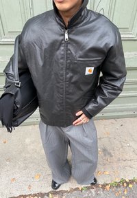 Melnas ādas žakete ar rāvējslēdzēju un augstu apkakli, ar krūšu kabatu un logotipa uzlīmi, kombinēta ar pelēkām plisētām biksēm un melnu somu.