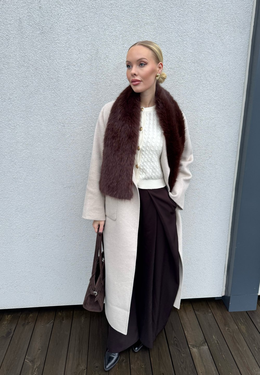 Helebeežist villane mantel pruuni karvase kraega, valge soonikkoes kampsun, tumedat pruunid laiade sääreosadega püksid ja tumedat pruunid käekott.