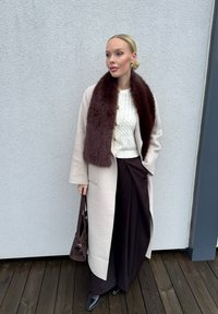 Helebeežist villane mantel pruuni karvase kraega, valge soonikkoes kampsun, tumedat pruunid laiade sääreosadega püksid ja tumedat pruunid käekott.