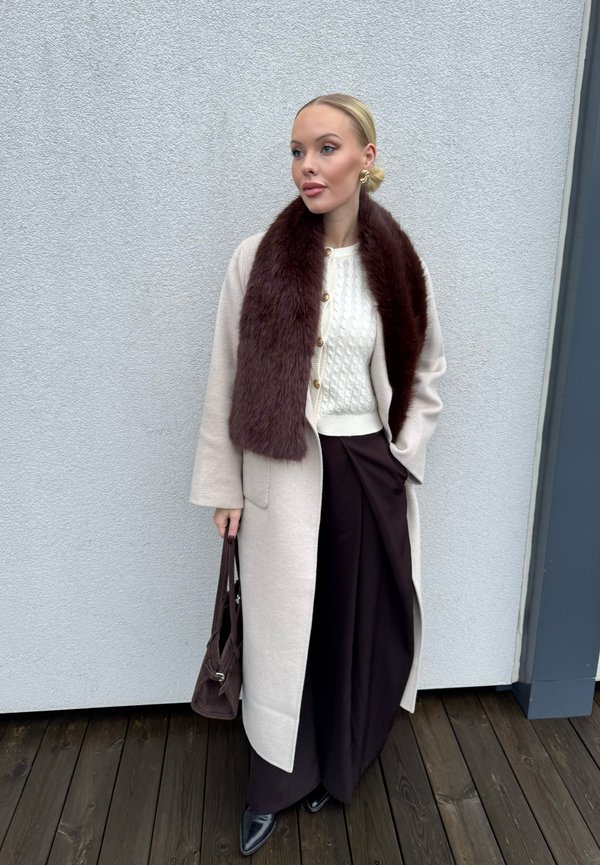 Ljusbeige ullrock med brun pälskrage, vit kabelstickad tröja, mörkbruna vida byxor och en mörkbrun handväska.