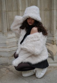 Plašč iz umetnega krzna v bledo rožnati barvi, z grobim dizajnom, kombiniran s črno mini obleko in prozornimi črnimi nogavicami. Celoten videz dopolnijo beli čevlji z vezalkami.