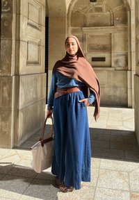 Lunga vestito blu con vita arricciata, cintura marrone e hijab marrone. Porta una borsa a spalla chiara, in piedi in un corridoio di pietra.