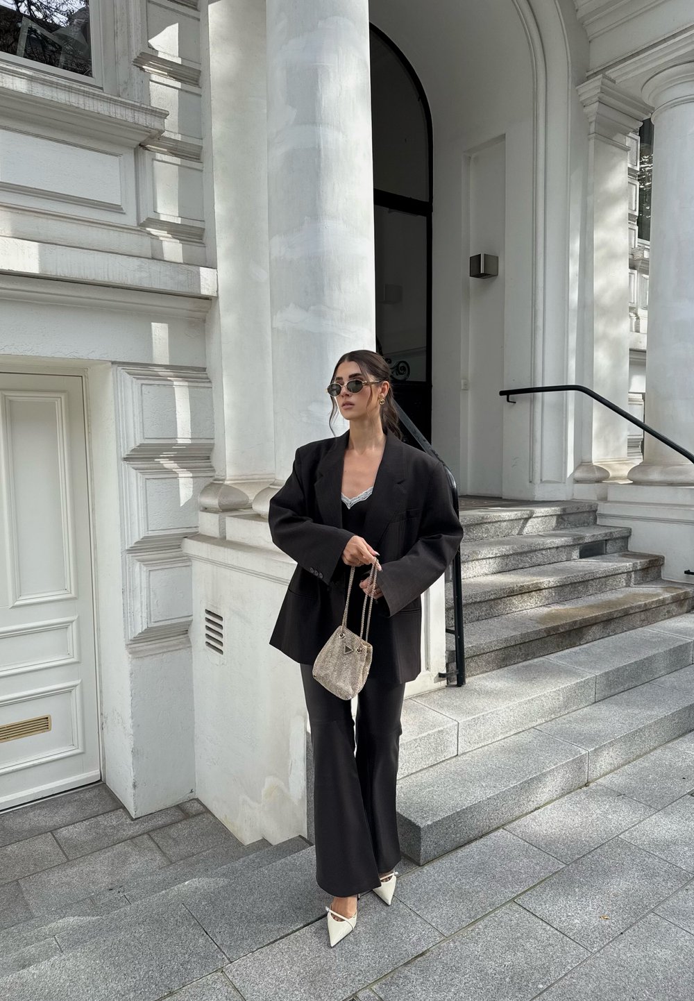 Zwarte oversized blazer over een zwart topje, gecombineerd met flared broeken en witte puntige hakken. Geaccessoireerd met een kleine geweven tas en een zonnebril.