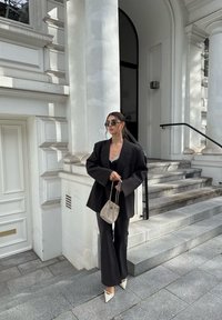 Zwarte oversized blazer over een zwart topje, gecombineerd met flared broeken en witte puntige hakken. Geaccessoireerd met een kleine geweven tas en een zonnebril.