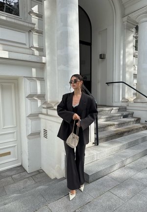 Čierny oversized blazer cez čierny top, doplnený o rozšírené nohavice a biele lodičky s špicatou špičkou. Ozdobené malou tkanou taškou a slnečnými okuliarmi.