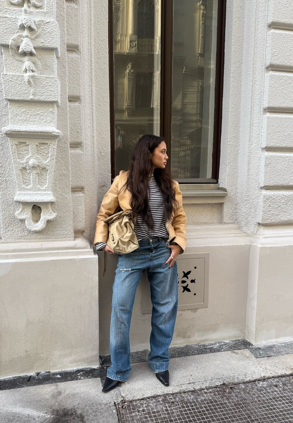 Hellbrauner Blazer über einem schwarz-weiß gestreiften Hemd, kombiniert mit weitem blauen Jeans und schwarzen spitzen Pumps. Beigefarbene Tasche über der Schulter getragen.