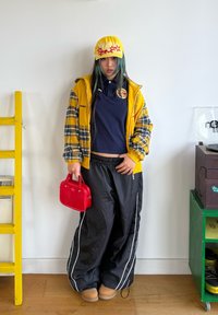 Gul caps med røde stjerner, rutete gul og blå jakke, mørk poloshirt, sorte vindbukser med hvite striper, rød håndveske, beige støvler.