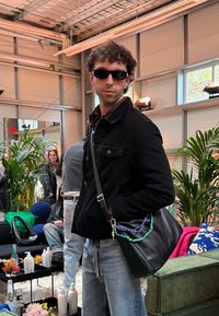 Giacca di jeans nera, jeans azzurri chiari, borsa nera con dettagli della catena colorata. Ambientazione interna con vegetazione e arredi morbidi sullo sfondo.