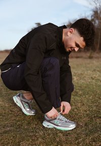 Silver athletic shoes with green accents, a mesh upper, and a cushioned sole. Black windbreaker and navy trousers; grassy outdoor setting.