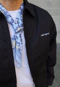 Schwarze Jacke mit silbernem Reißverschluss und Logo, weißes Hemd darunter und ein blau gemusterter Schal um den Hals gebunden.
