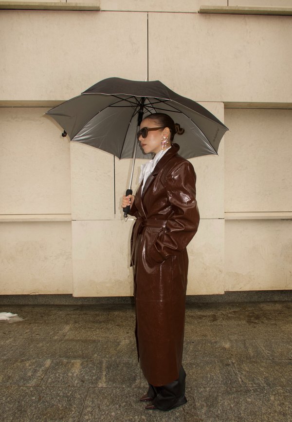 Frau in braunem Leder-Trenchcoat hält einen schwarz-silbernen Regenschirm, trägt Sonnenbrille und große, bunte Ohrringe, steht auf einem Steinpflaster.