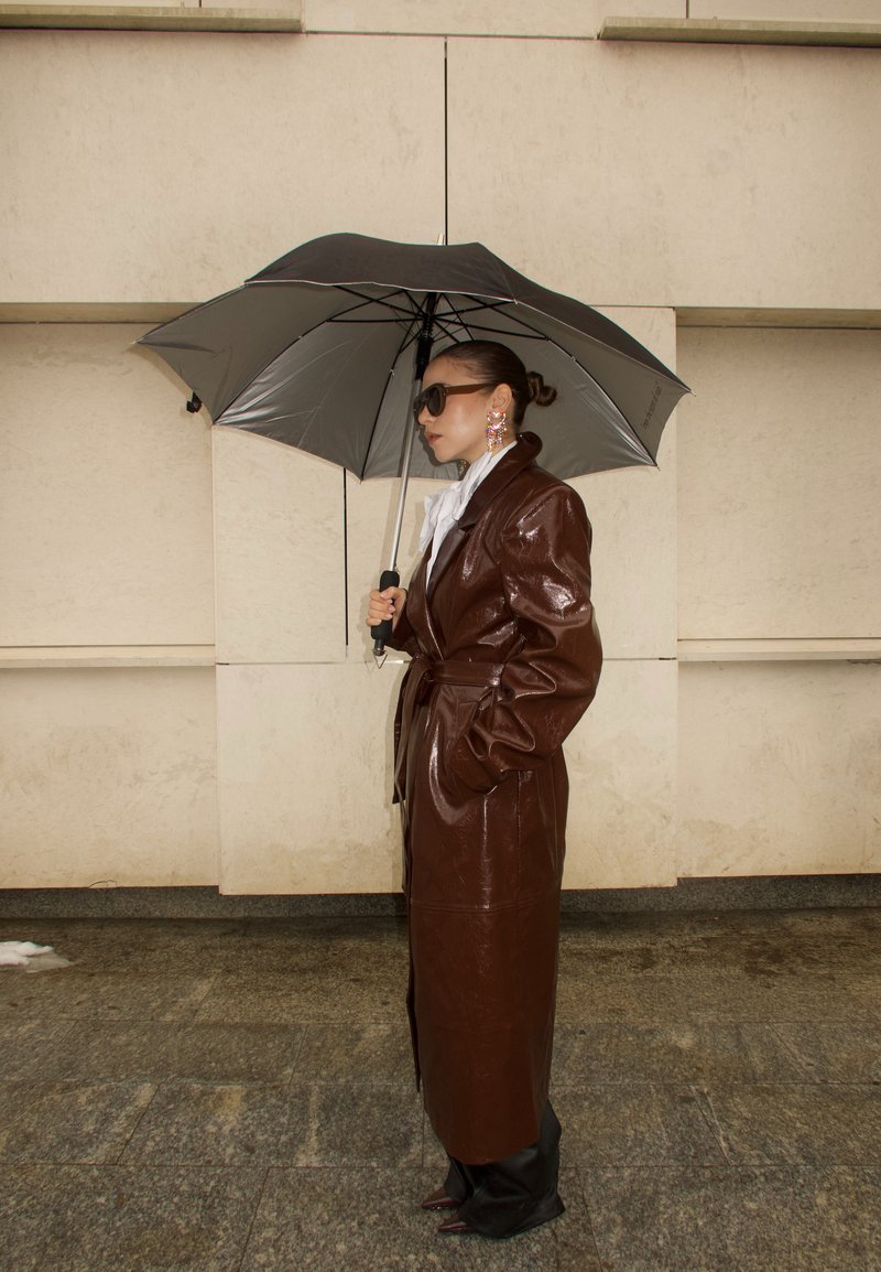 Donna con trench in pelle marrone che tiene un ombrello nero e argento, indossa occhiali da sole e grandi orecchini colorati, in piedi su un pavimento in pietra.