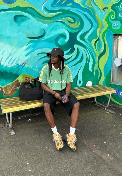 Jersey verde de Adidas con cuello blanco, pantalones cortos negros, zapatillas amarillas, sombrero tipo cubo negro y gafas, sentado en un banco amarillo frente a un mural colorido.