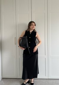 Musta hihaton toppi kultaisilla napeilla, yhdistettynä liehuvaan mustaan hameeseen. Mustaa käsilaukkua pidetään käsivarren alla, taustalla vaaleaa kaappia.