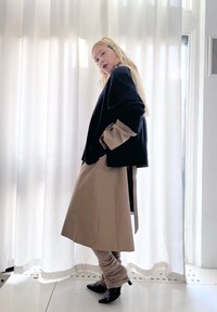 Woman standing sideways wearing a black jumper over a beige coat, knitted trousers, black pointed shoes, and hair clips, with hands in pockets.